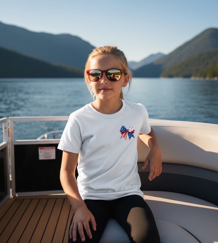 JSD Apparel youth white t-shirt with patriotic bow graphic, worn by a girl on a boat during a summer day.