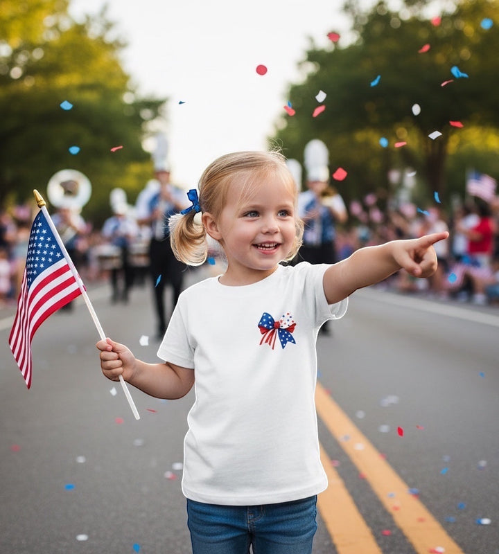 JSD Apparel youth tee with patriotic bow graphic, worn by a girl holding an American flag at a parade.