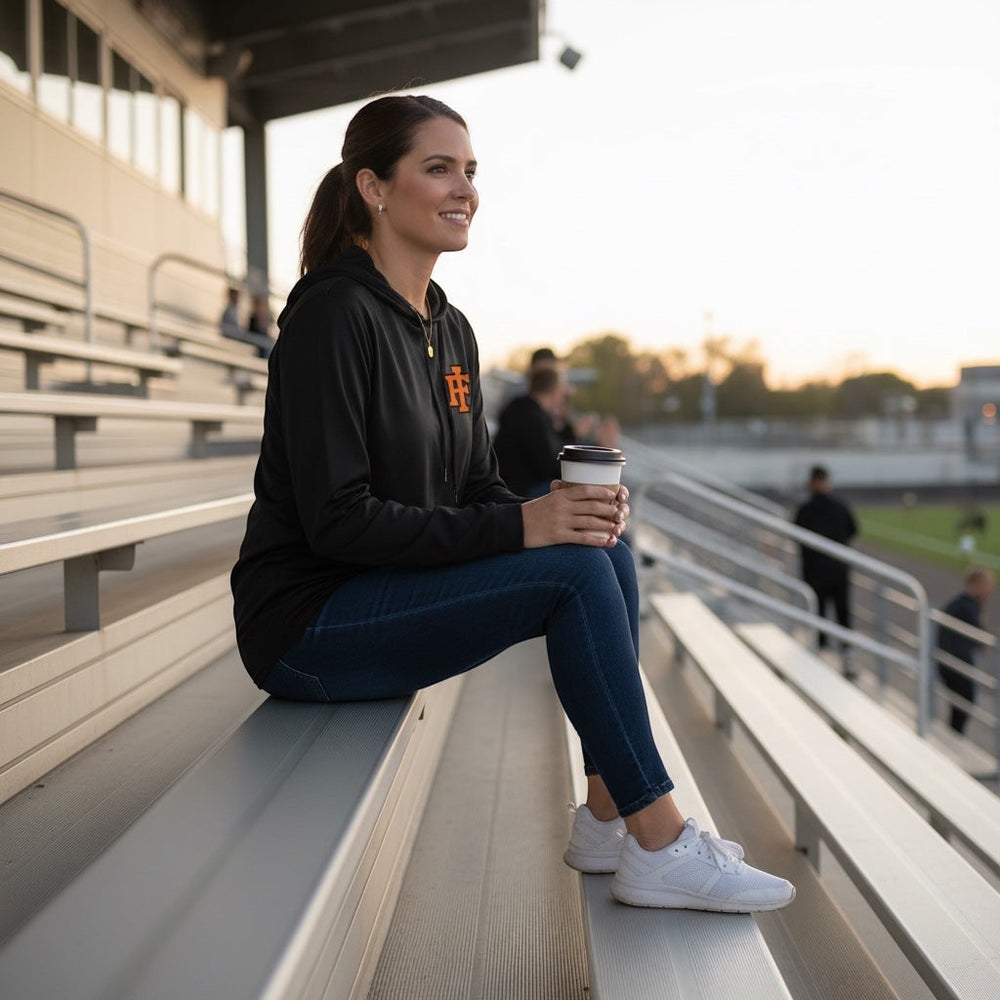A woman wearing a black JSD Apparel performance hooded long sleeve with an orange IF logo, sitting on stadium bleachers.