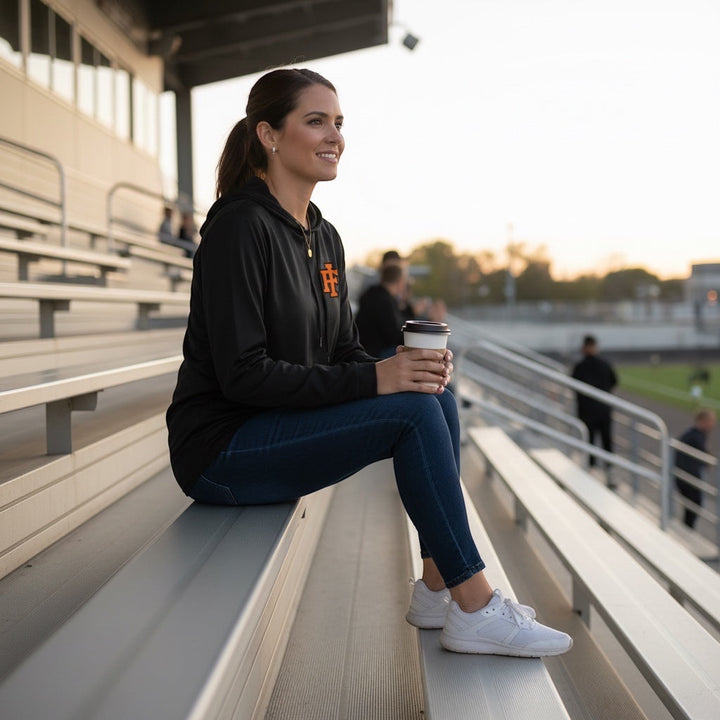 A woman wearing a black JSD Apparel performance hooded long sleeve with an orange IF logo, sitting on stadium bleachers.