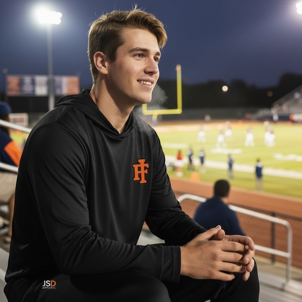 A man wearing a black JSD Apparel performance hooded long sleeve with an orange IF logo at a night football game.