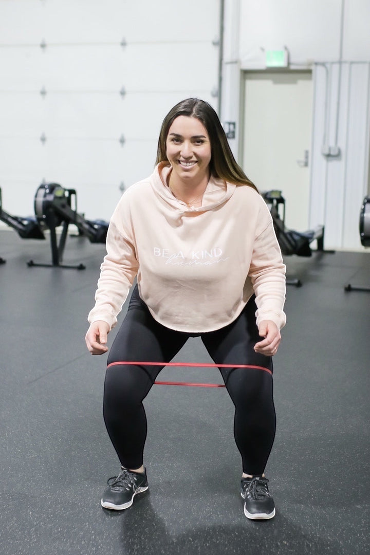 JSD Apparel light pink cropped hooded sweatshirt with Be A Kind Human text worn by a woman doing squats in a gym