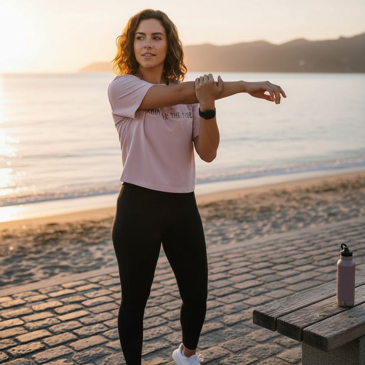 JSD Apparel light pink cropped t-shirt with "Train for the Tides" graphic, worn by a woman stretching on a beach.