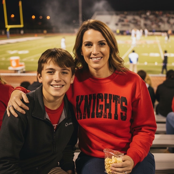 Woman wearing a red JSD Apparel Hillcrest Knights vintage arch crewneck sweatshirt at a night football game
