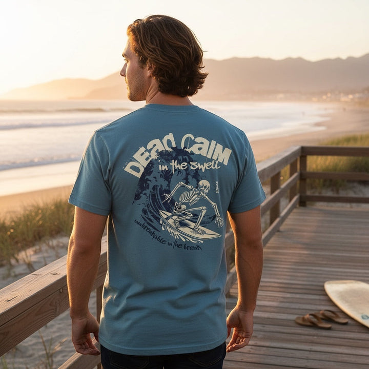 Man wearing JSD Apparel blue cotton t-shirt with skeleton surfer back graphic at a beach at sunset.
