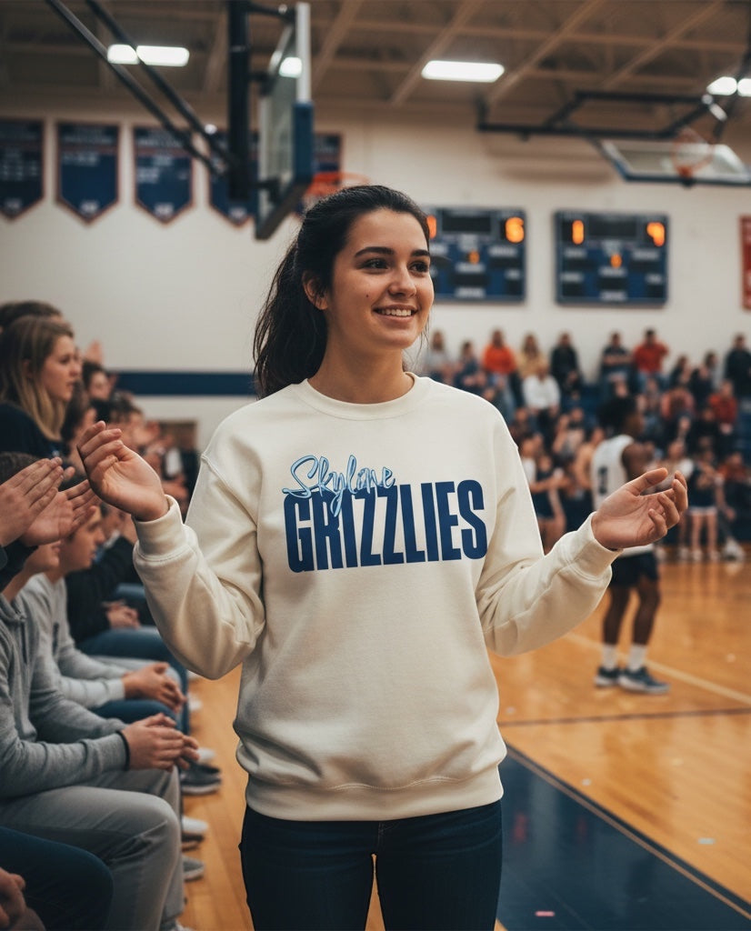 Woman wearing a cream JSD Apparel Skyline Grizzlies crewneck sweatshirt with blue script lettering at a basketball game