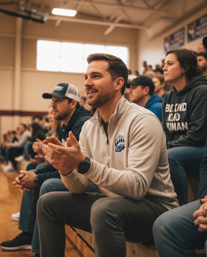 Man wearing a JSD Apparel Skyline Grizzlies quarter zip pullover with a paw logo, sitting in a crowded sports arena.