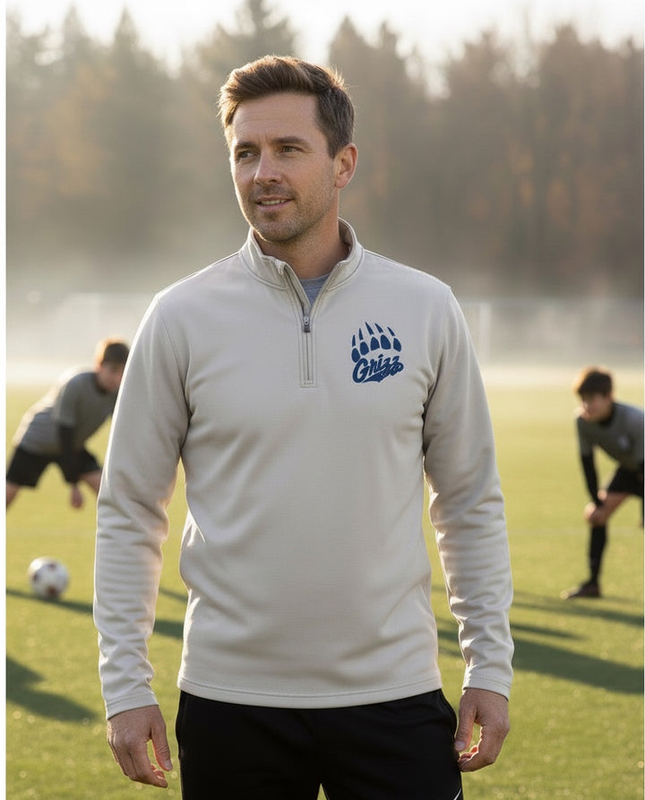 Man wearing a beige JSD Apparel Skyline Grizzlies quarter zip pullover with a blue paw logo on a sports field.