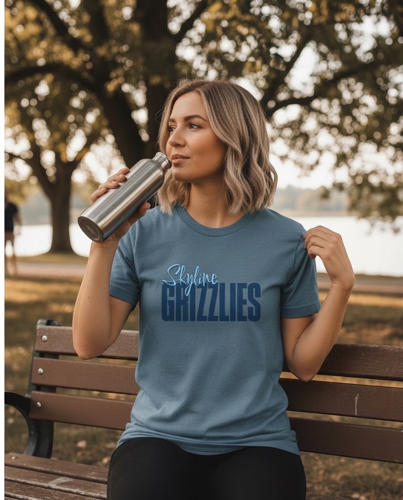 Woman wearing a blue JSD Apparel Skyline Grizzlies cotton tee while drinking from a metal bottle on a park bench.