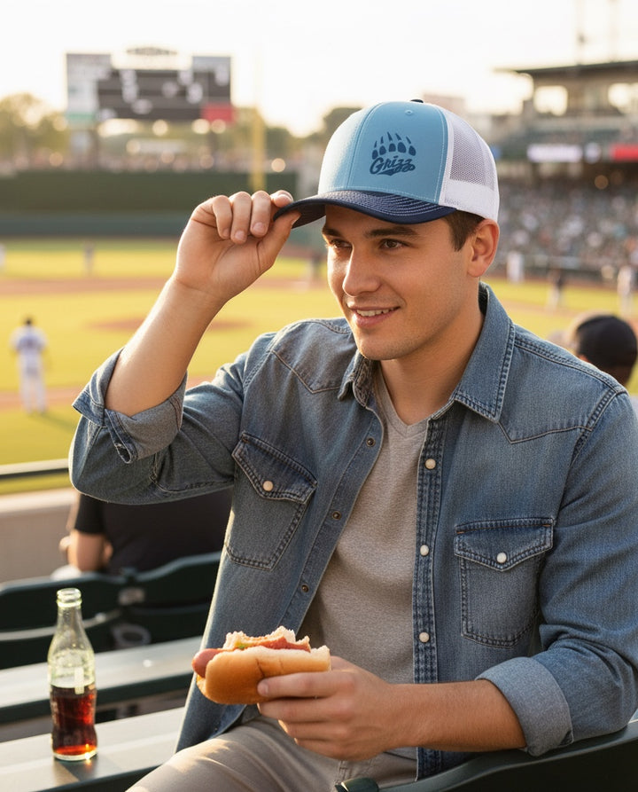 JSD Apparel Skyline Grizzlies paw trucker hat in light blue and white worn by a man at a baseball stadium.