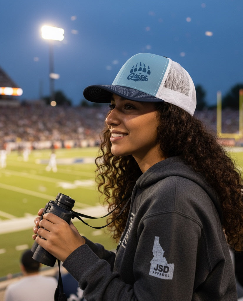 Woman wearing a JSD Apparel Skyline Grizzlies paw trucker hat with a light blue front and white mesh back at a stadium.