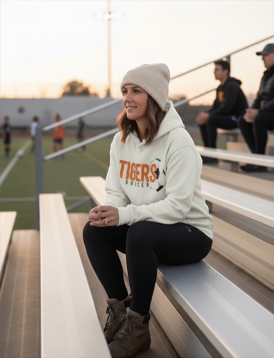 JSD Apparel Tiger Pride soccer hoodie in cream with orange distressed graphic, worn by a woman on stadium bleachers.