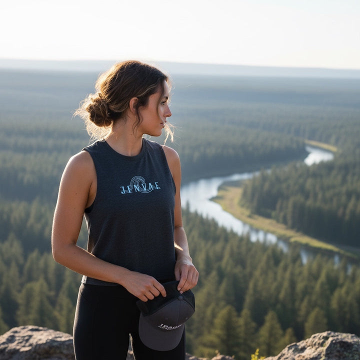 Woman wearing a JSD Apparel deep charcoal heather cropped tank with a wave logo, standing outdoors overlooking a river.