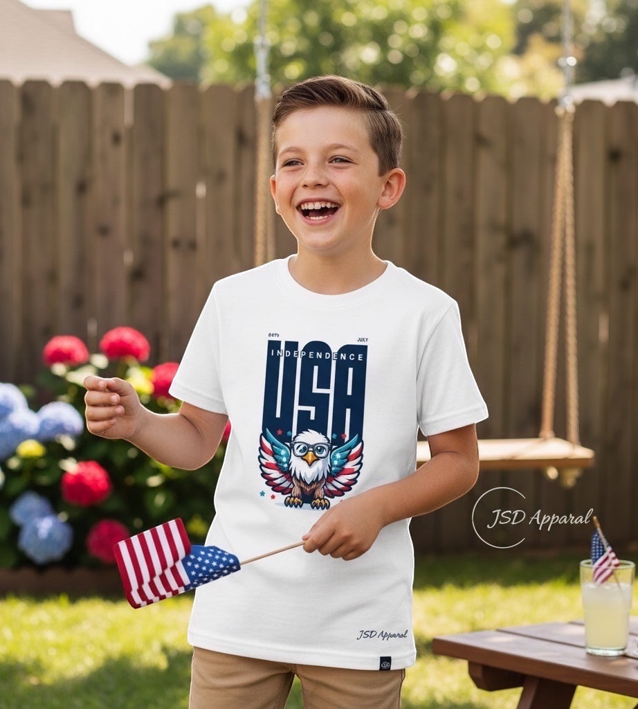 A smiling boy wearing a JSD Apparel white youth tee with a patriotic eagle graphic, holding a small American flag.